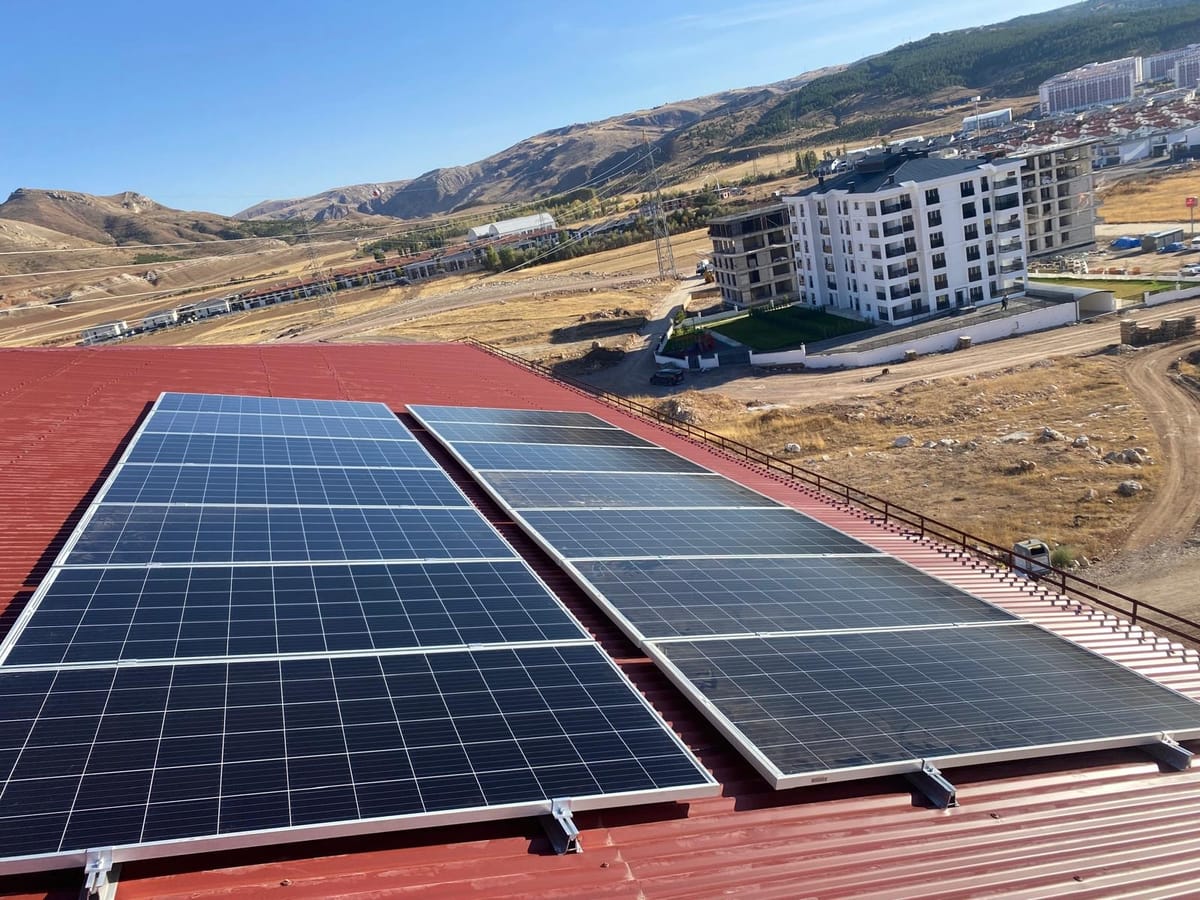 Tepelere bakan bir işletmenin kırmızı sac çatısına iki sıra hâlinde kurulan güneş panelleri